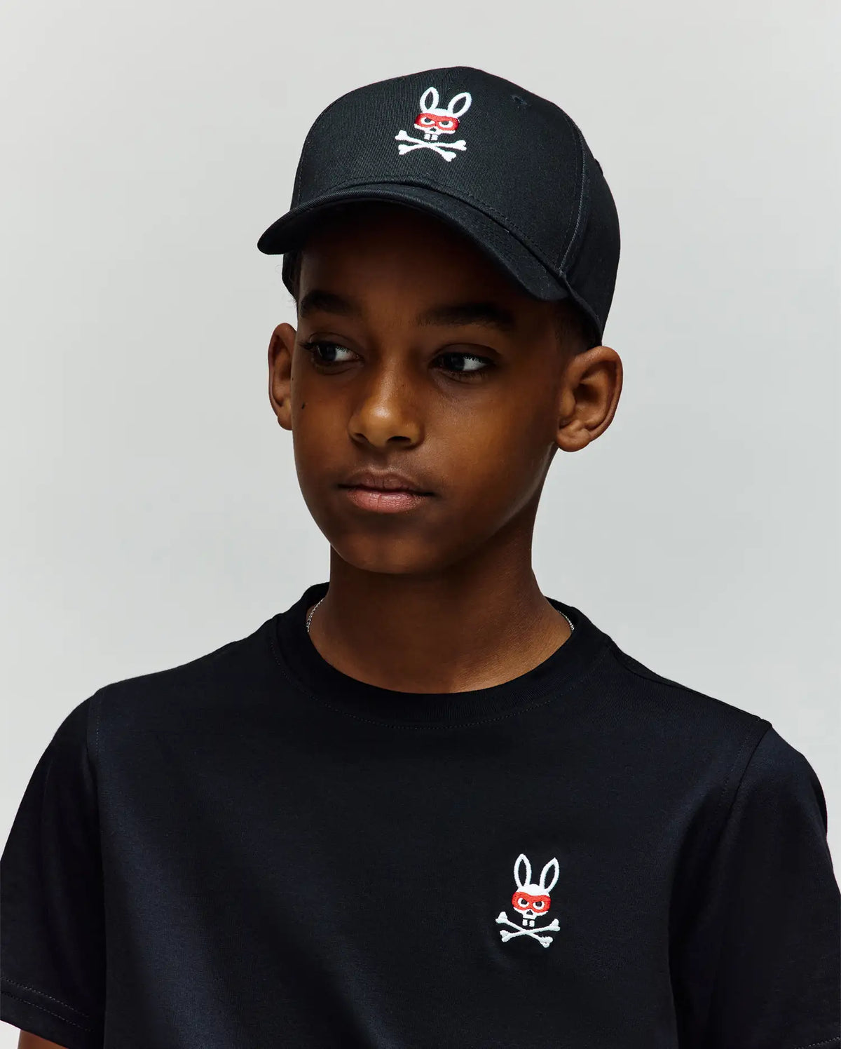 A boy stands against a light gray background, wearing a 100% cotton black t-shirt and the Psycho Bunny KIDS MISCHIEF BASEBALL CAP (KO1500013), both featuring the white and red embroidered Mischief Bunny skull-and-crossbones logo.