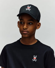 A boy stands against a light gray background, wearing a 100% cotton black t-shirt and the Psycho Bunny KIDS MISCHIEF BASEBALL CAP (KO1500013), both featuring the white and red embroidered Mischief Bunny skull-and-crossbones logo.