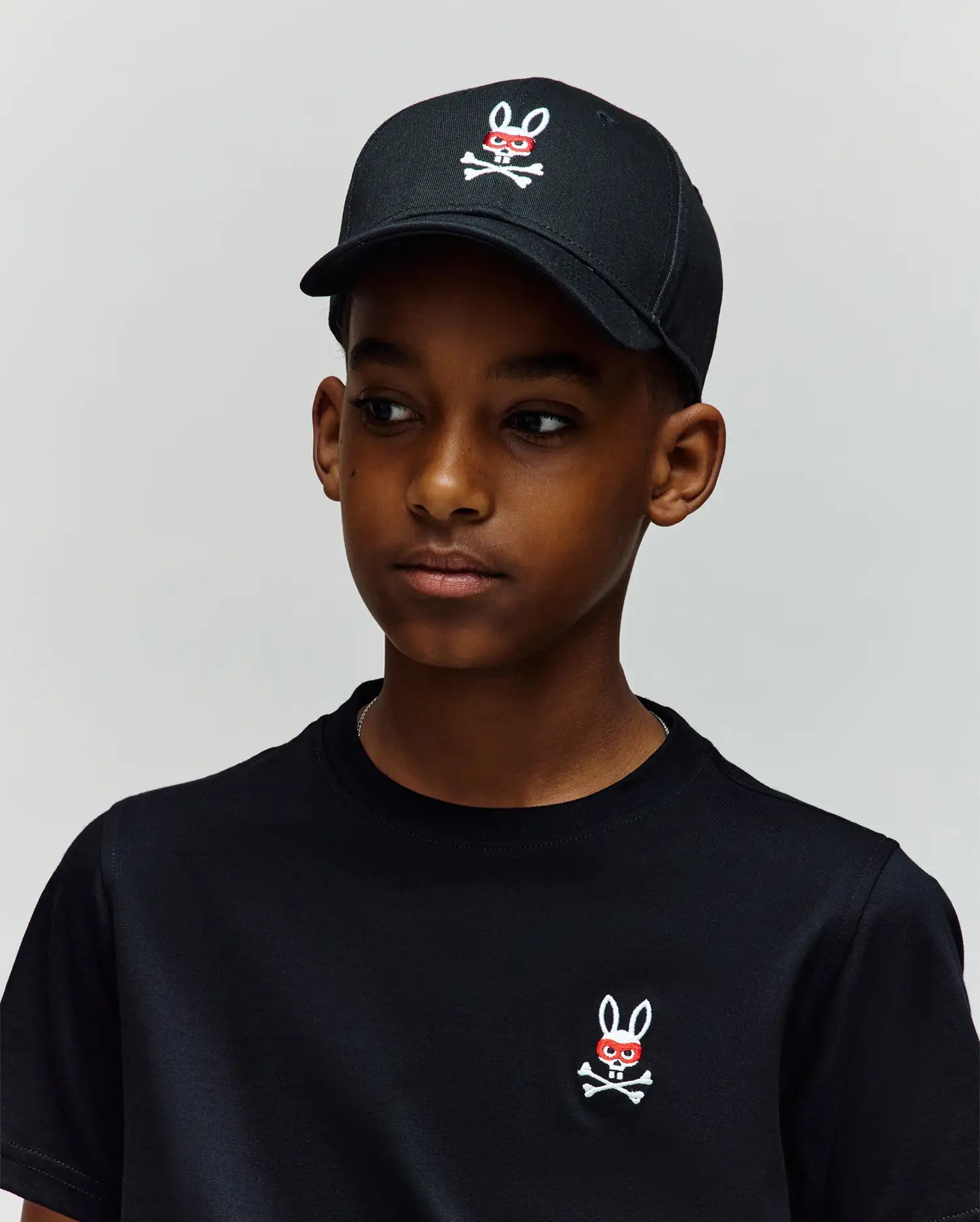 A boy stands against a light gray background, wearing a 100% cotton black t-shirt and the Psycho Bunny KIDS MISCHIEF BASEBALL CAP (KO1500013), both featuring the white and red embroidered Mischief Bunny skull-and-crossbones logo.