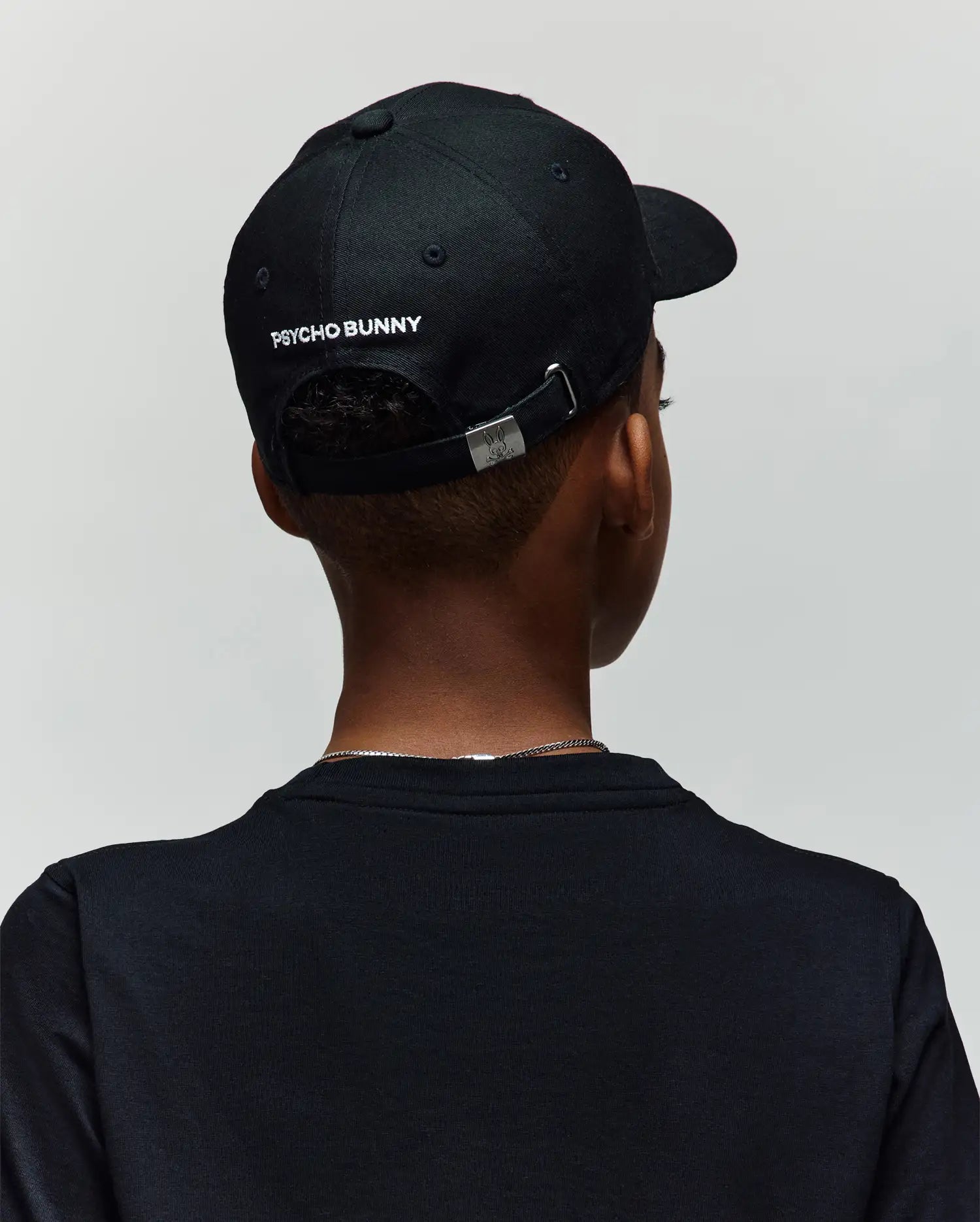 A person wearing a black Psycho Bunny KIDS MISCHIEF BASEBALL CAP (KO1500013) with an embroidered Mischief Bunny on the back, facing away from the camera. The 100% cotton cap is paired with a black shirt and thin chain necklace; background is light.