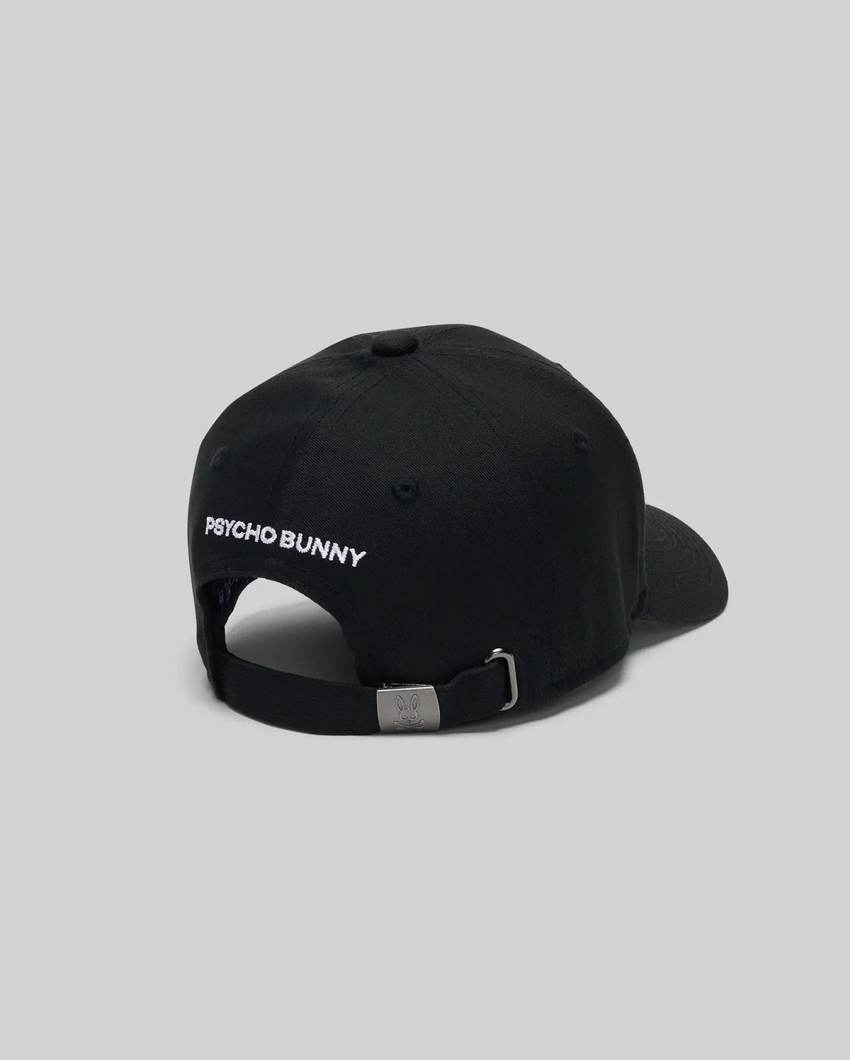 The KIDS MISCHIEF BASEBALL CAP - KO1500013 by Psycho Bunny is a black 100% cotton cap with "PSYCHO BUNNY" embroidered in white on the back, featuring an adjustable metal buckle. Displayed against a plain gray background.
