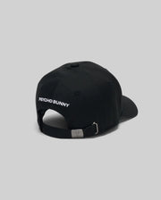 The KIDS MISCHIEF BASEBALL CAP - KO1500013 by Psycho Bunny is a black 100% cotton cap with "PSYCHO BUNNY" embroidered in white on the back, featuring an adjustable metal buckle. Displayed against a plain gray background.