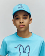 A young person wears the Psycho Bunny KIDS CYRUS BASEBALL CAP (KO1500014) and matching t-shirt, both with the brand’s bunny logo, standing against a plain light background and looking directly at the camera with a neutral expression.