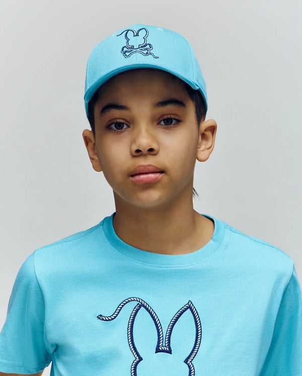 A young person wears the Psycho Bunny KIDS CYRUS BASEBALL CAP (KO1500014) and matching t-shirt, both with the brand’s bunny logo, standing against a plain light background and looking directly at the camera with a neutral expression.