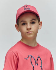 A young person wears the Psycho Bunny KIDS CYRUS BASEBALL CAP (KO1500014), a pink cotton twill hat, and a pink t-shirt with the brand's bunny logo, standing against a plain light background and looking at the camera.