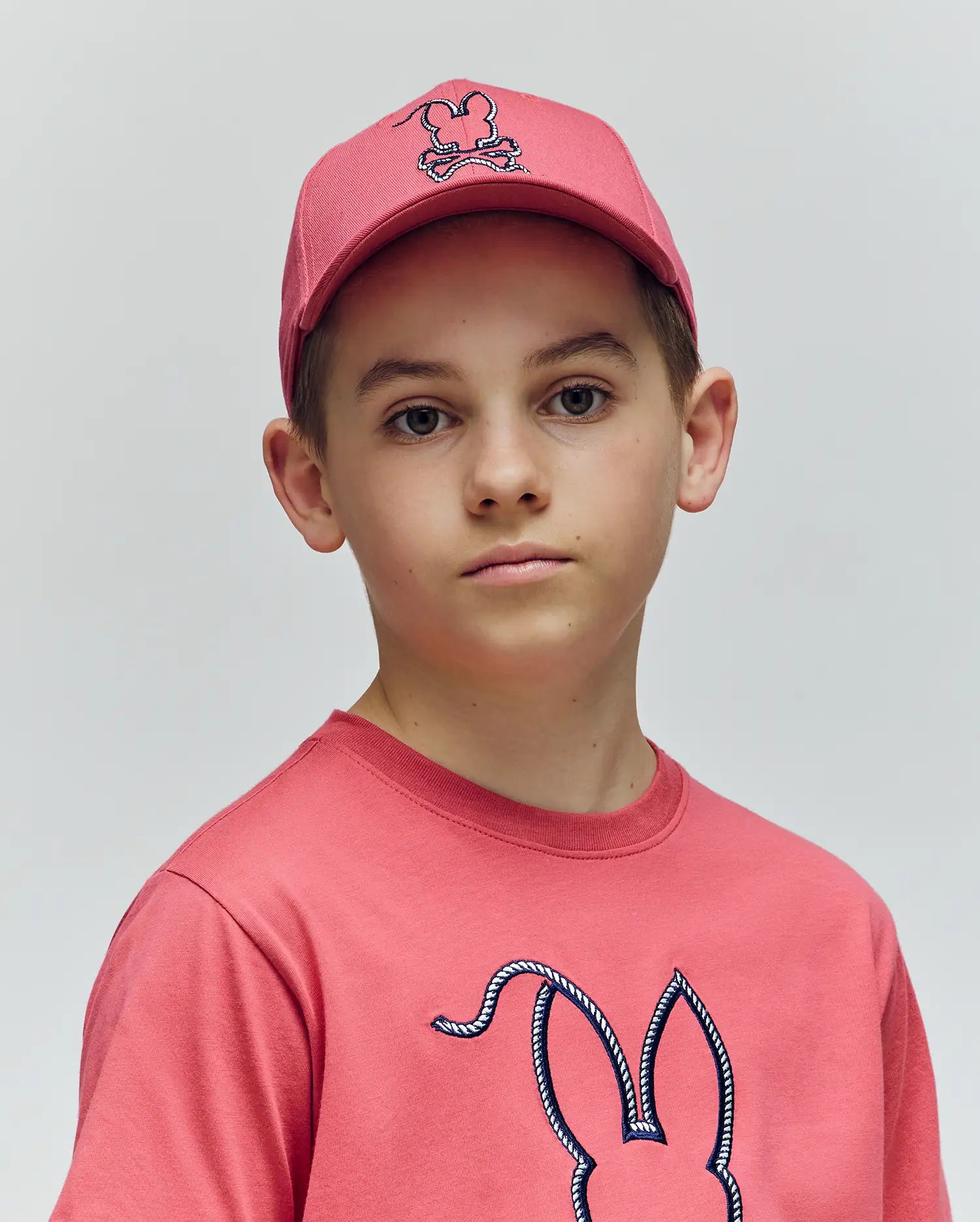 A young person wears the Psycho Bunny KIDS CYRUS BASEBALL CAP (KO1500014), a pink cotton twill hat, and a pink t-shirt with the brand's bunny logo, standing against a plain light background and looking at the camera.