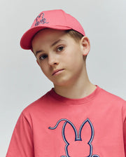 A boy wears the Psycho Bunny KIDS CYRUS BASEBALL CAP (KO1500014) and a matching pink tee with a bunny graphic, looking at the camera with a neutral expression against a plain light background.