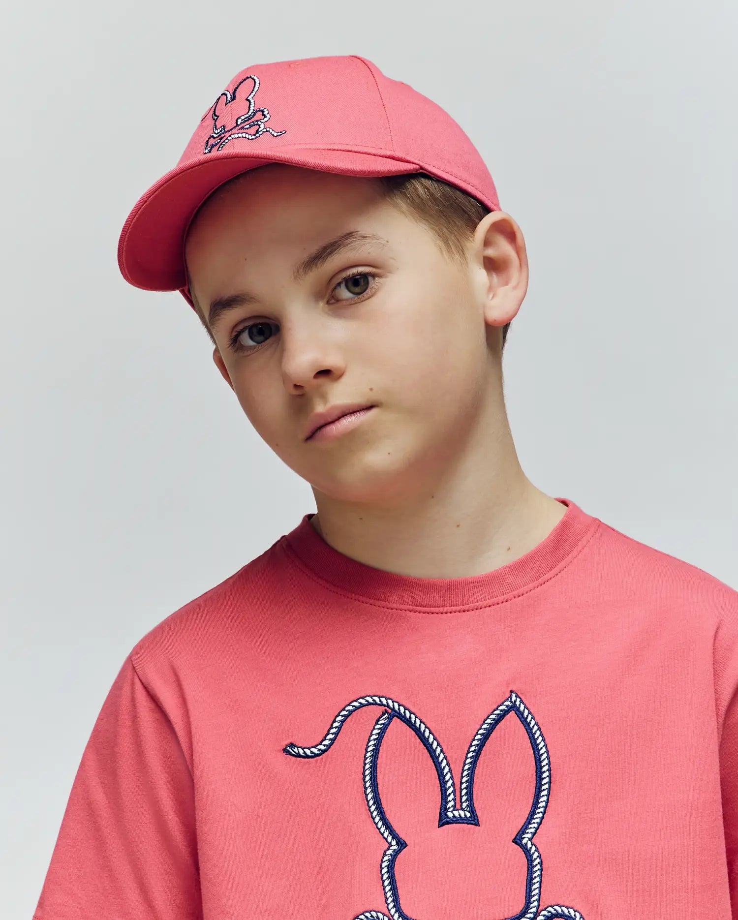 A boy wears the Psycho Bunny KIDS CYRUS BASEBALL CAP (KO1500014) and a matching pink tee with a bunny graphic, looking at the camera with a neutral expression against a plain light background.