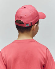 A young boy is seen from behind wearing a pink Psycho Bunny KIDS CYRUS BASEBALL CAP (KO1500014) with embroidered initials and an adjustable strap, paired with a matching t-shirt against a plain light background.