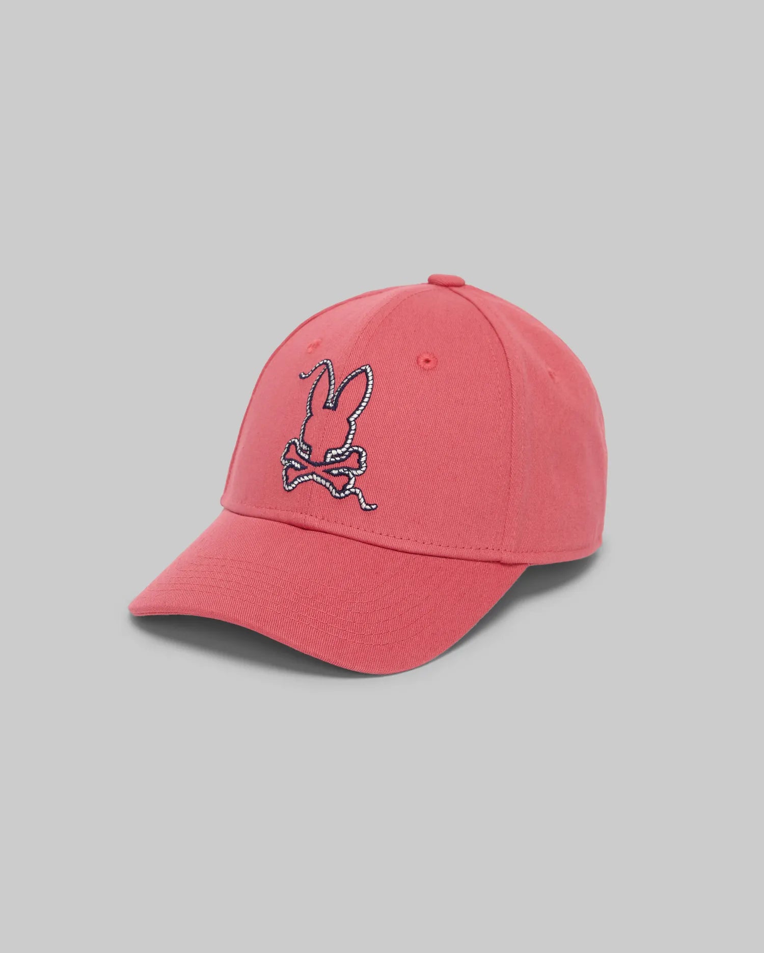 The Psycho Bunny KIDS CYRUS BASEBALL CAP (KO1500014) is a pink cotton twill hat with an embroidered bunny head and crossed bones on the front, shown on a light gray background. This adjustable cap combines comfort with playful style.
