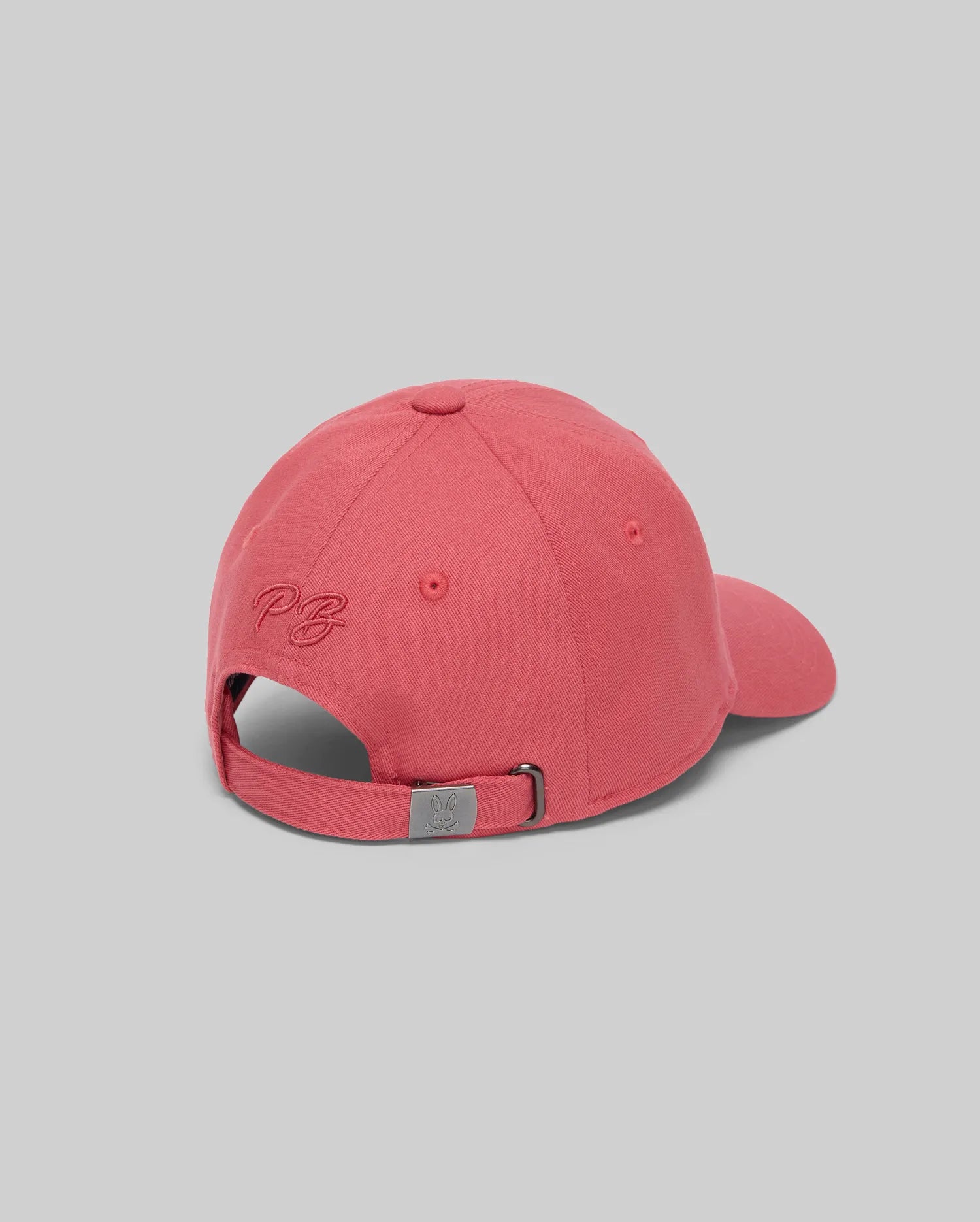 The Psycho Bunny KIDS CYRUS BASEBALL CAP (KO1500014) is a pink cotton twill adjustable hat seen from the back, with a metal clasp, subtle "PB" embroidery above the opening, set against a plain light grey background.