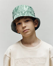 A young boy with curly hair wears the Psycho Bunny KIDS CHUCK REVERSIBLE BUCKET HAT (KO1500015) and a cream sweatshirt with embossed text, standing against a plain light background.