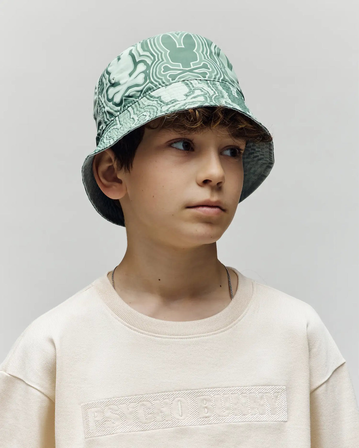 A young boy with wavy brown hair wears the Psycho Bunny KIDS CHUCK REVERSIBLE BUCKET HAT (KO1500015) in a mint green pattern and a cream sweatshirt, looking to the side with a neutral expression against a plain background.