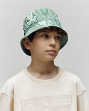 A young boy with wavy brown hair wears the Psycho Bunny KIDS CHUCK REVERSIBLE BUCKET HAT (KO1500015) in a mint green pattern and a cream sweatshirt, looking to the side with a neutral expression against a plain background.