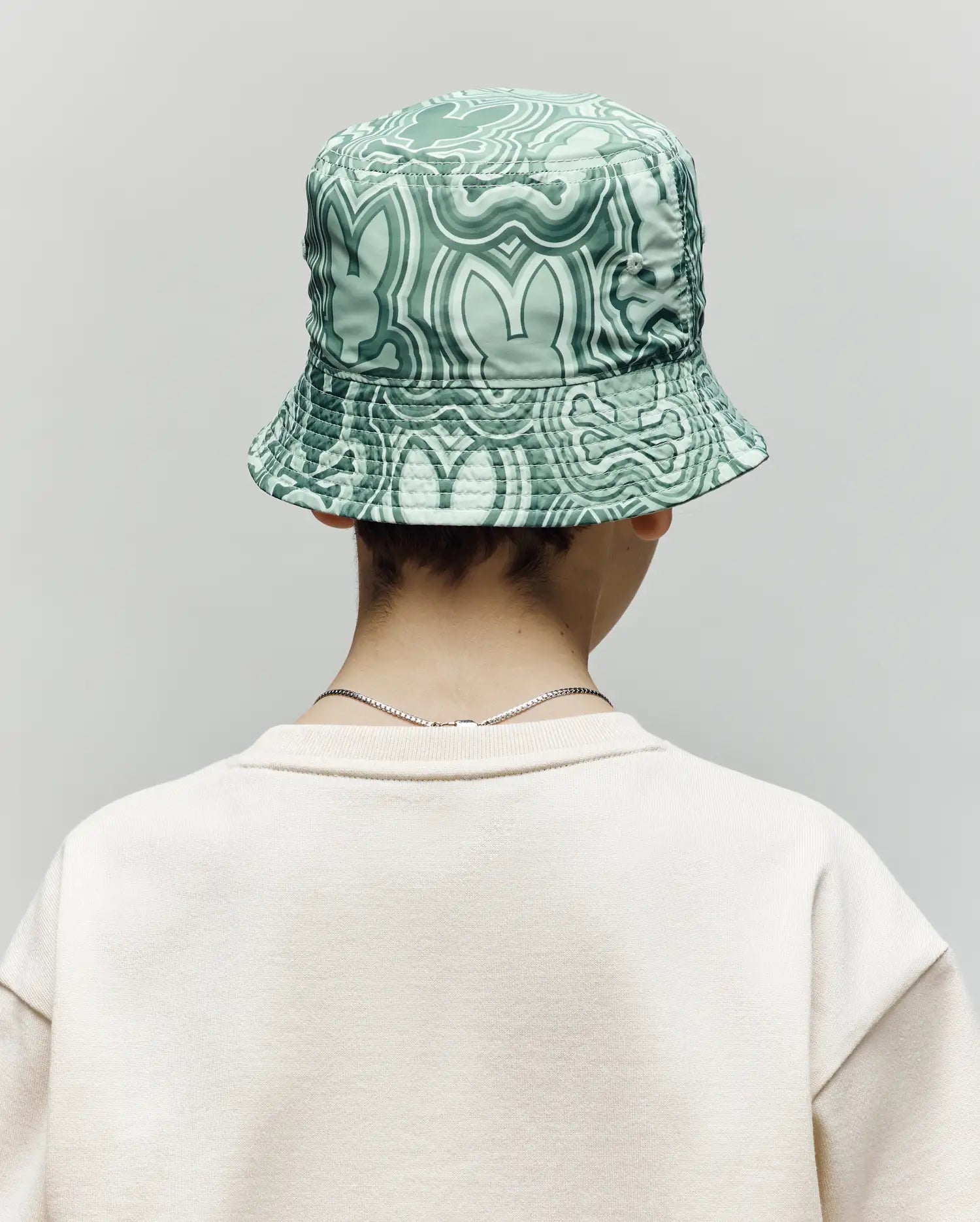 A person is seen from behind in a light beige sweatshirt and the Psycho Bunny KIDS CHUCK REVERSIBLE BUCKET HAT (KO1500015), which features a green pattern and SPF protection, against a plain gray background.