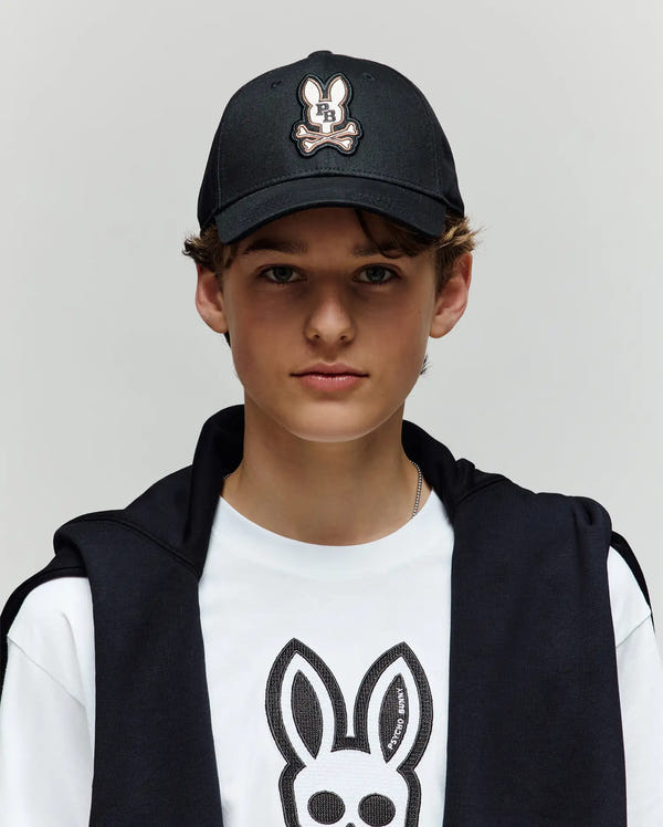 A young person with light skin and brown hair wears the Psycho Bunny KIDS WADE BASEBALL CAP (KO1500016), featuring an embroidered Bunny patch, along with a matching white t-shirt and a black jacket, against a plain background.