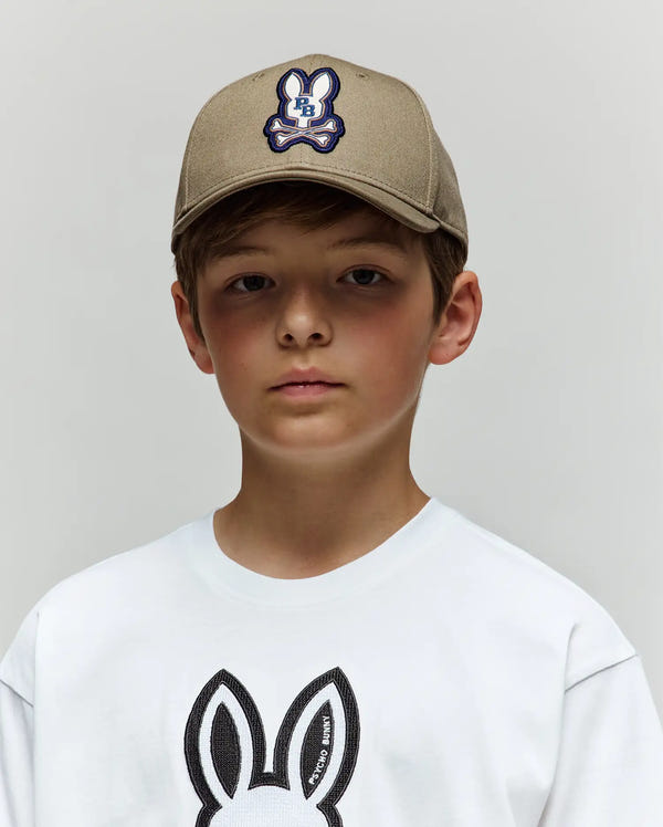 A young boy in a beige Psycho Bunny KIDS WADE BASEBALL CAP (KO1500016) with an embroidered bunny logo and a white bunny graphic T-shirt stands against a plain light gray background.