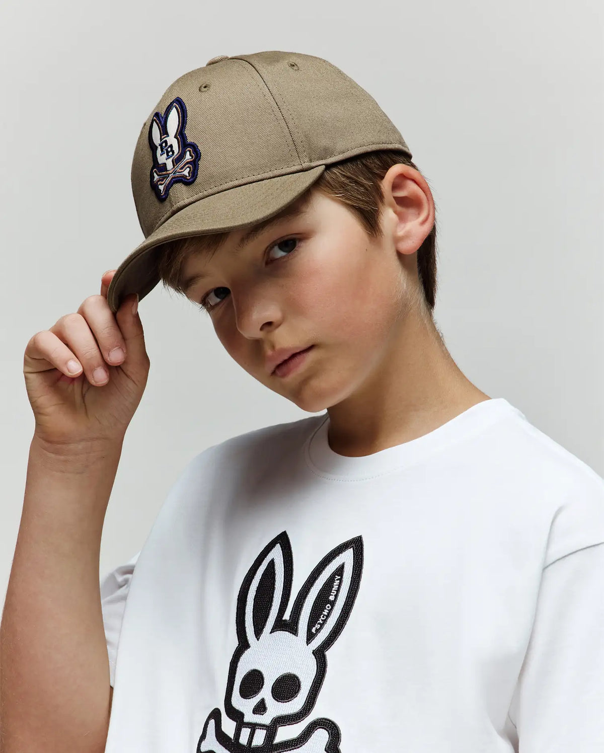 A young boy in a Psycho Bunny KIDS WADE BASEBALL CAP (KO1500016) with a tan cotton twill finish and embroidered Bunny logo, plus a white tee with a large black bunny skull graphic, tilts his cap and looks at the camera against a plain backdrop.