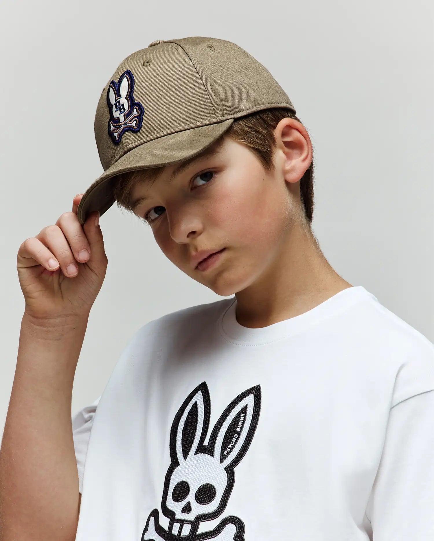 A young boy in a Psycho Bunny KIDS WADE BASEBALL CAP (KO1500016), crafted from beige cotton twill with an embroidered Bunny skull logo, and a matching graphic tee, looks at the camera holding his cap’s brim against a plain gray background.