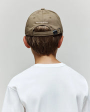 A child wears the Psycho Bunny KIDS WADE BASEBALL CAP (KO1500016) in light brown cotton twill, featuring an embroidered Bunny and "Psycho Bunny" text on the back, paired with a white T-shirt, shown from behind against a plain gray background.