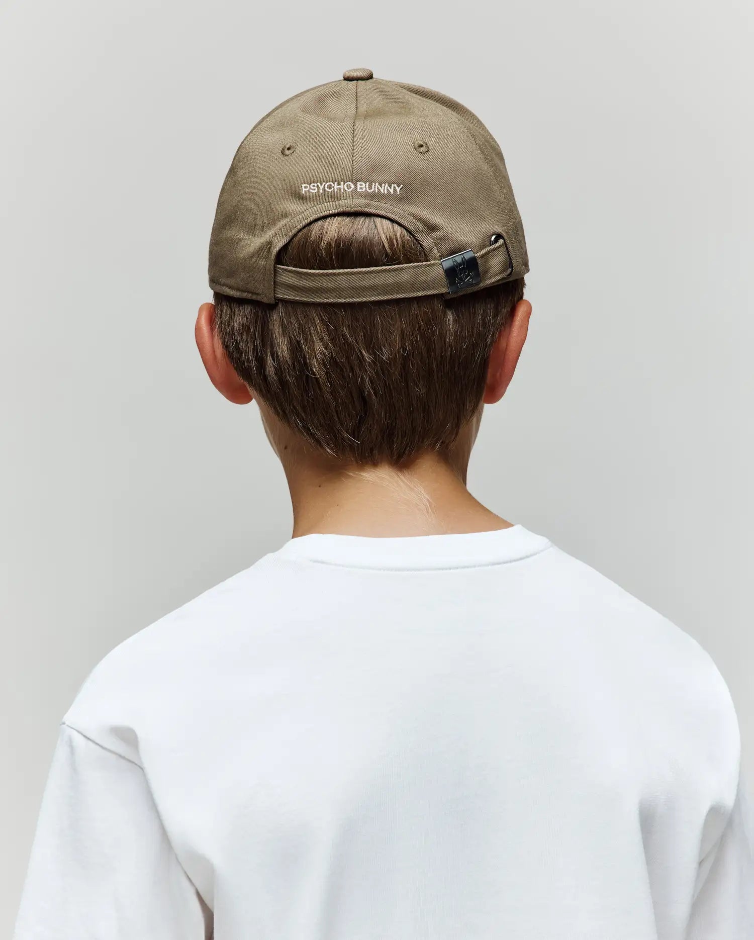 A child wears the Psycho Bunny KIDS WADE BASEBALL CAP (KO1500016) in light brown cotton twill, featuring an embroidered Bunny and "Psycho Bunny" text on the back, paired with a white T-shirt, shown from behind against a plain gray background.