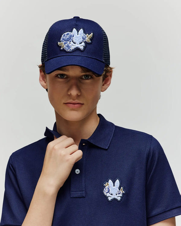 A young person wears the Psycho Bunny KIDS AUGUSTE TRUCKER CAP - KO1500022, a navy blue snapback with a white and blue bunny patch, paired with a matching polo shirt, standing against a plain light background and looking directly at the camera.