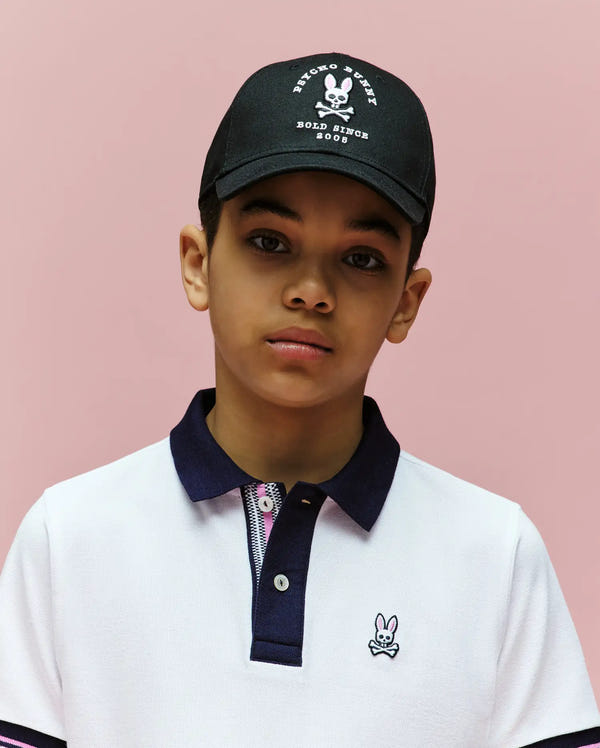 A young boy wears the Psycho Bunny KIDS ARCHIVE BASEBALL CAP - KO1500004 in black with a white graphic, paired with a white polo featuring a navy collar and embroidered bunny logo, posing against a plain pink background.
