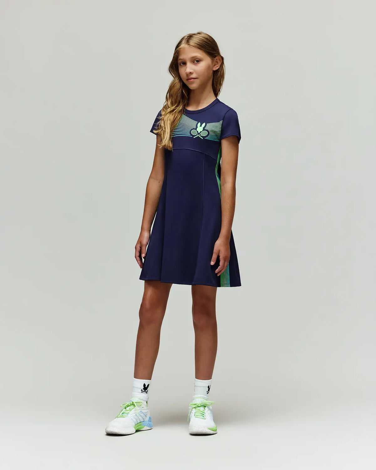 A young girl stands against a plain light background, wearing the Psycho Bunny RETAIL BALL CREW GIRLS DRESS (FR) - KO2000004, paired with white socks featuring a black logo and white athletic shoes with green and blue details.