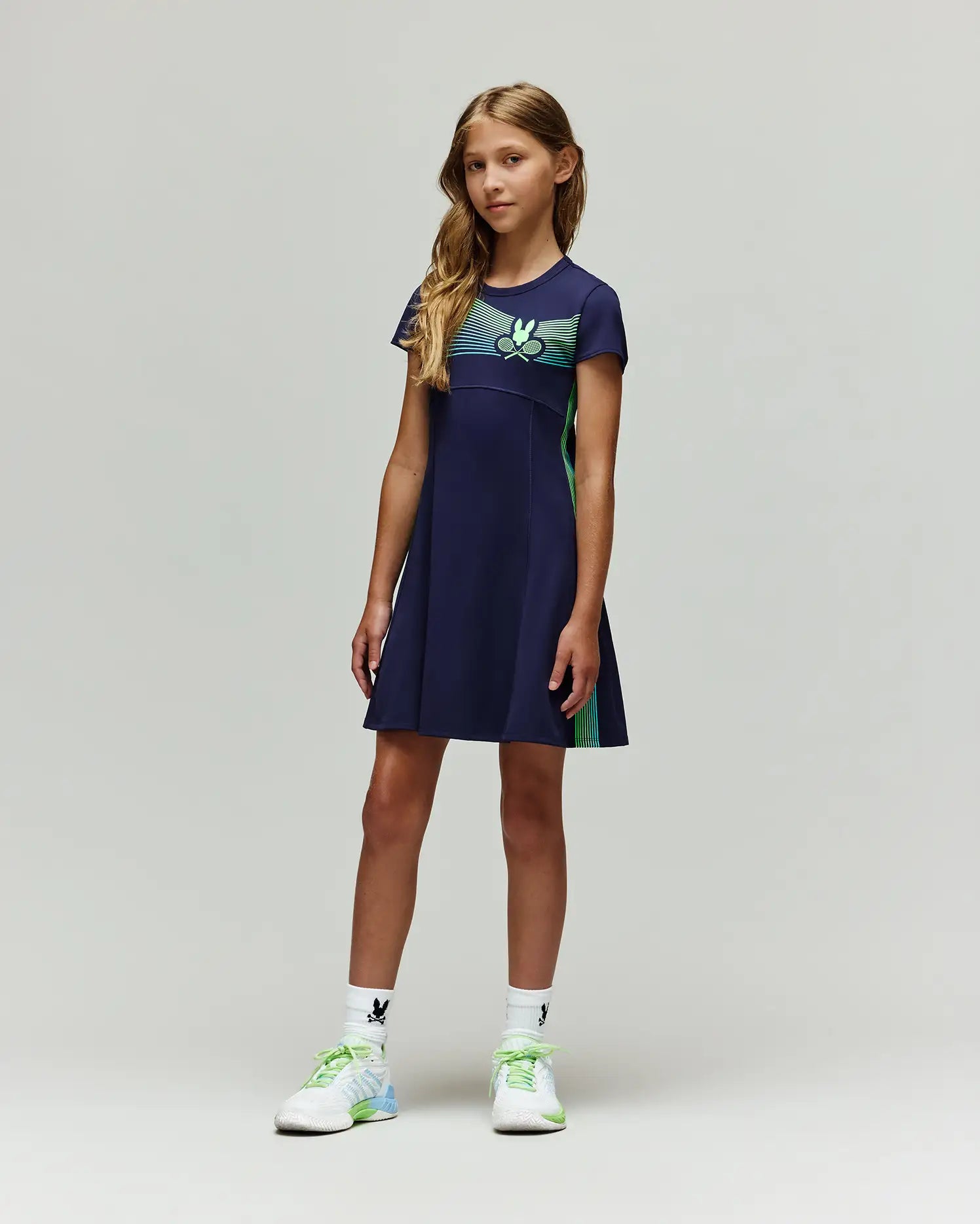 A young girl stands against a plain light background, wearing the Psycho Bunny RETAIL BALL CREW GIRLS DRESS (FR) - KO2000004, paired with white socks featuring a black logo and white athletic shoes with green and blue details.