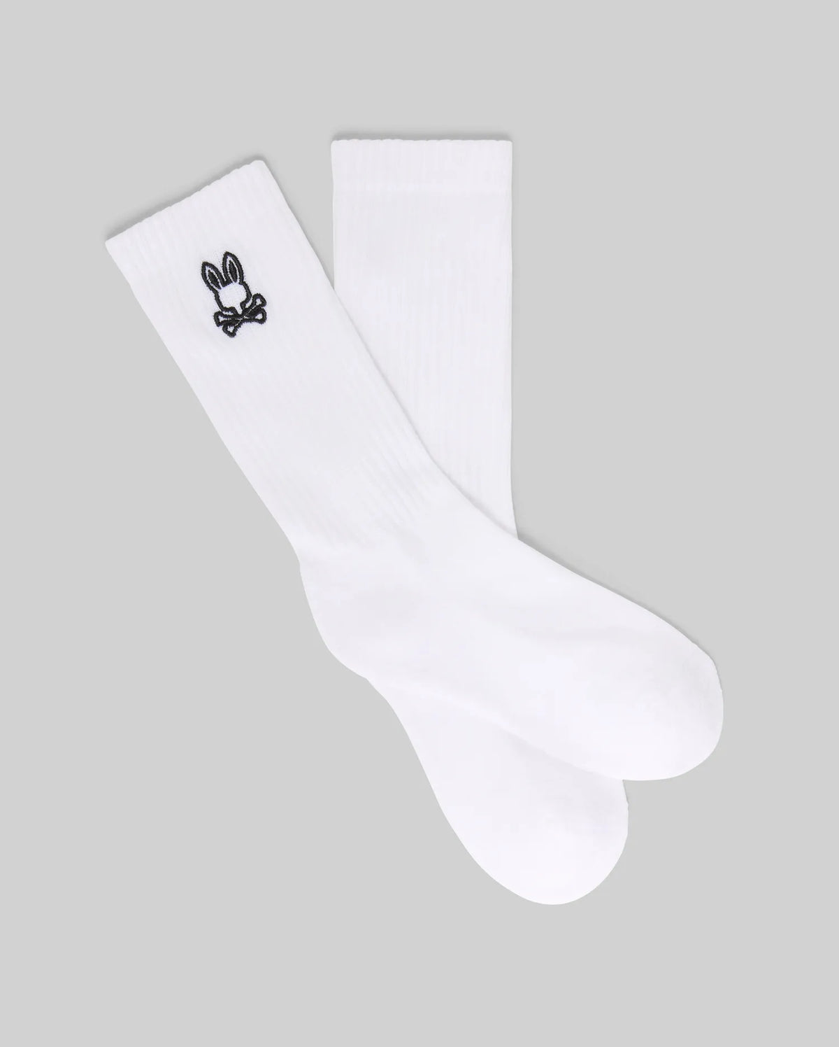 The Psycho Bunny KIDS 3 PACK SPORT SOCK (KO2200001) includes white crew socks made with soft Pima cotton and spandex, each featuring a small black bunny skull and crossbones logo near the top. Displayed on a gray background.