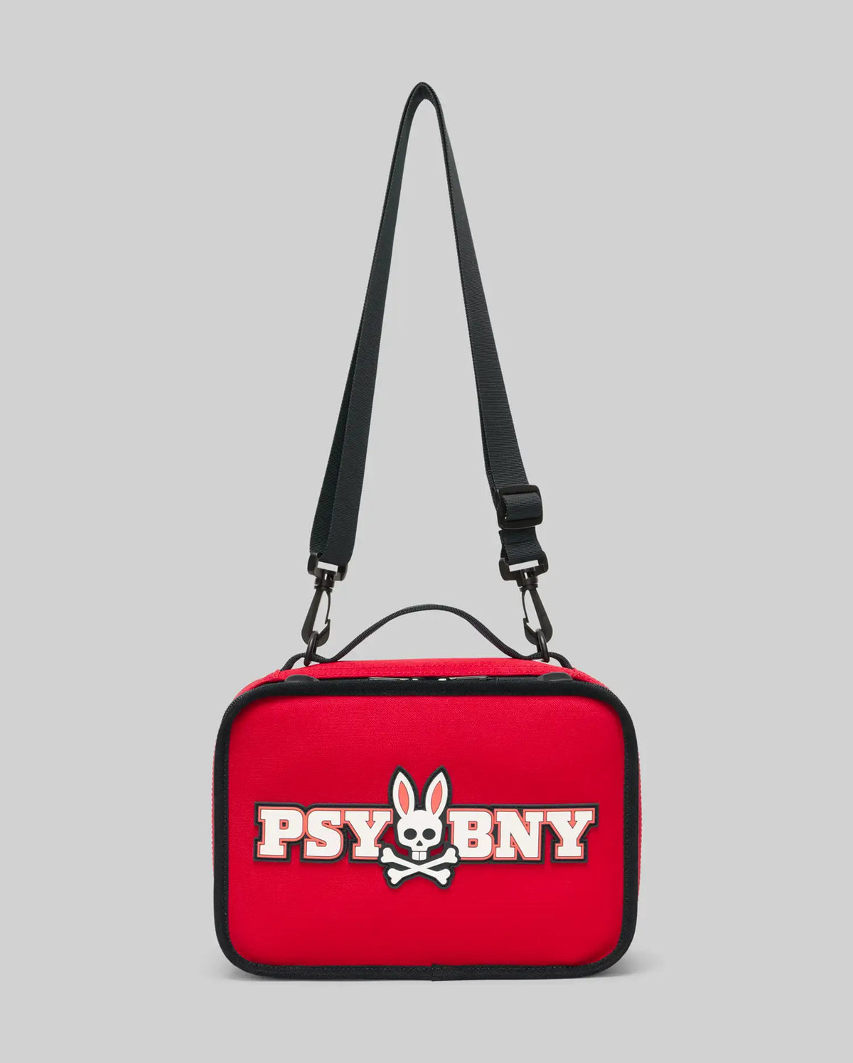 The PSYCHO BUNNY LUNCH BOX - KO2400002 by Psycho Bunny is a rectangular red insulated lunch box with an adjustable black strap, white bunny skull and crossbones logo, and "PSYBNY" text on the front. Background is light gray.
