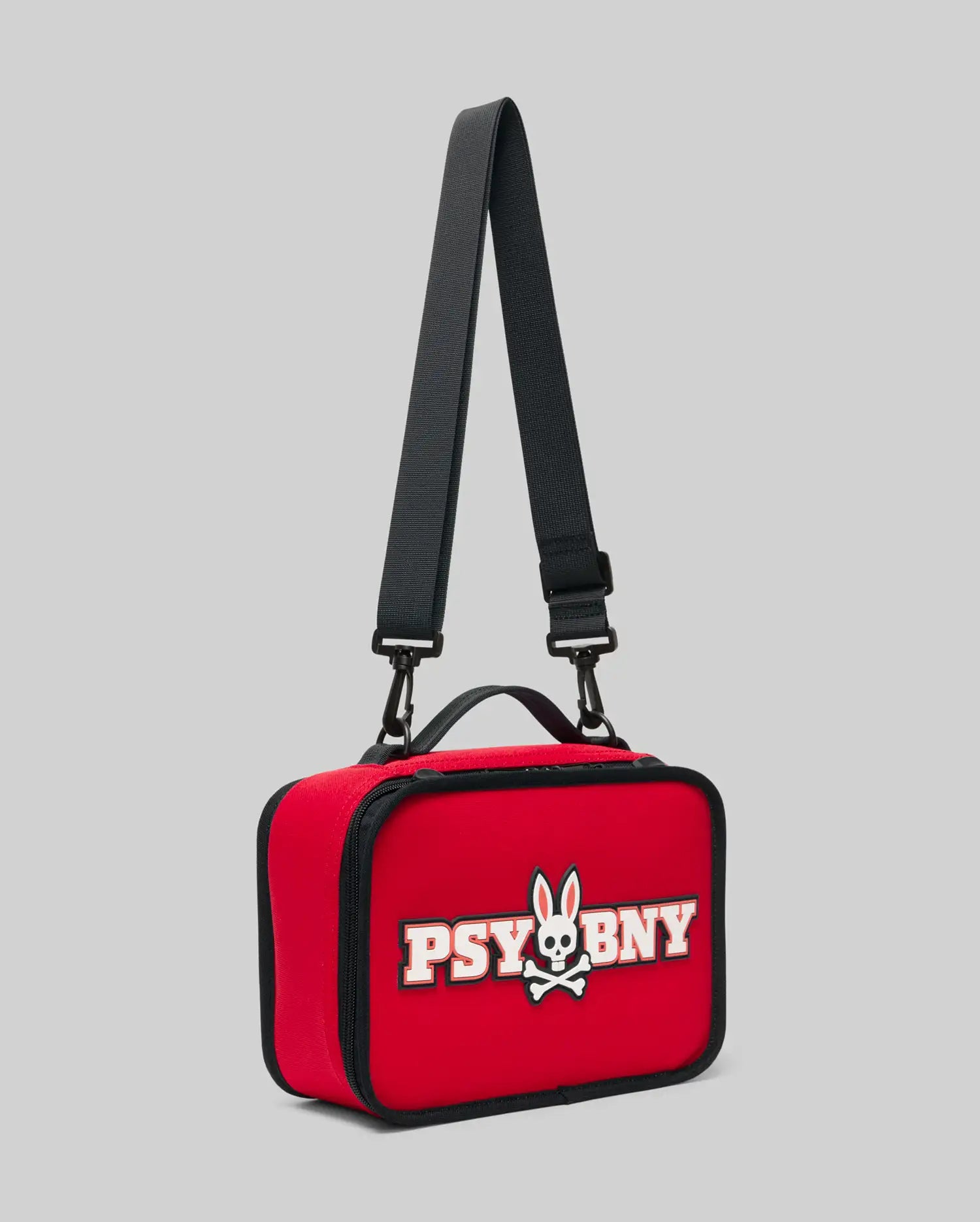The PSYCHO BUNNY LUNCH BOX - KO2400002 by Psycho Bunny is a red rectangular silicone bag with a black adjustable strap, black zipper, and a front patch showing a bunny skull and crossbones with 