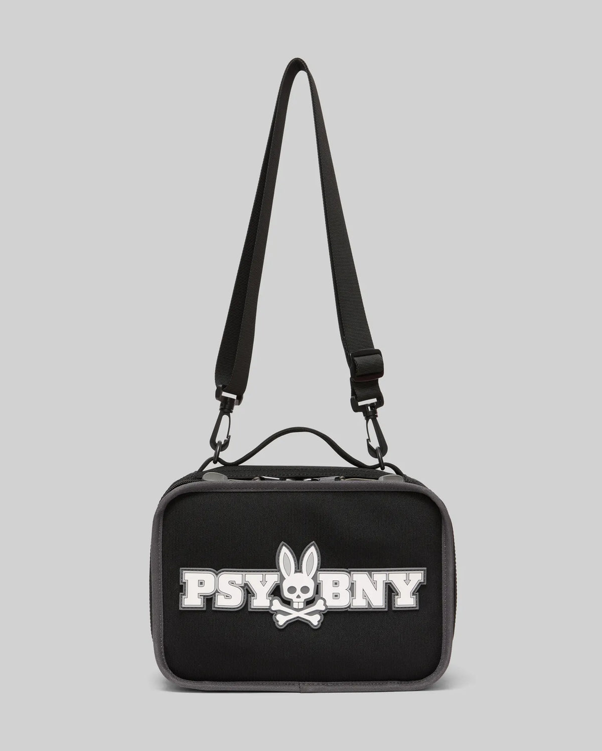 The Psycho Bunny Lunch Box (KO2400002) by Psycho Bunny is black with a top handle, adjustable shoulder strap, and features a white silicone Bunny with skull and crossbones plus "PSYBNY" on the front against a light gray background.