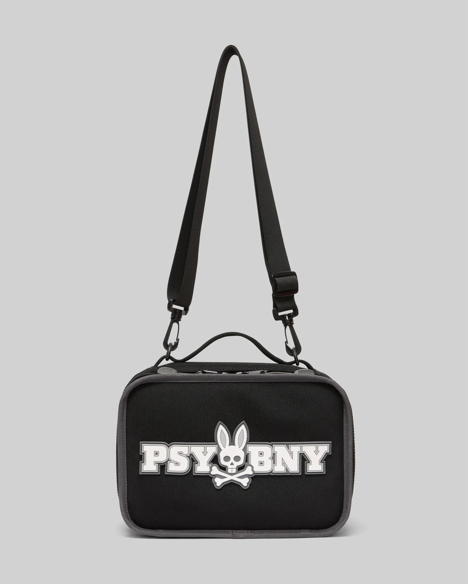 The Psycho Bunny Lunch Box (KO2400002) by Psycho Bunny is black with a top handle, adjustable shoulder strap, and features a white silicone Bunny with skull and crossbones plus "PSYBNY" on the front against a light gray background.