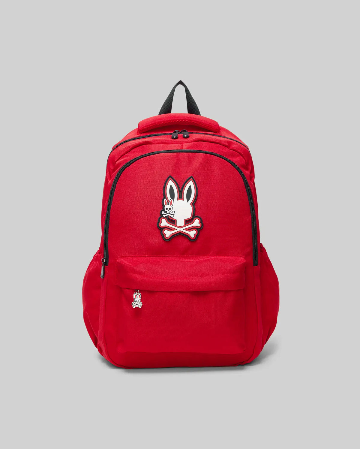The KIDS PSYCHO BUNNY BACKPACK - KO2400004 by Psycho Bunny is a bright red canvas kids' bag with padded straps, front pocket, zipper, cartoon bunny with crossbones logo above the pocket, and a matching pin. Shown on a plain light gray background.