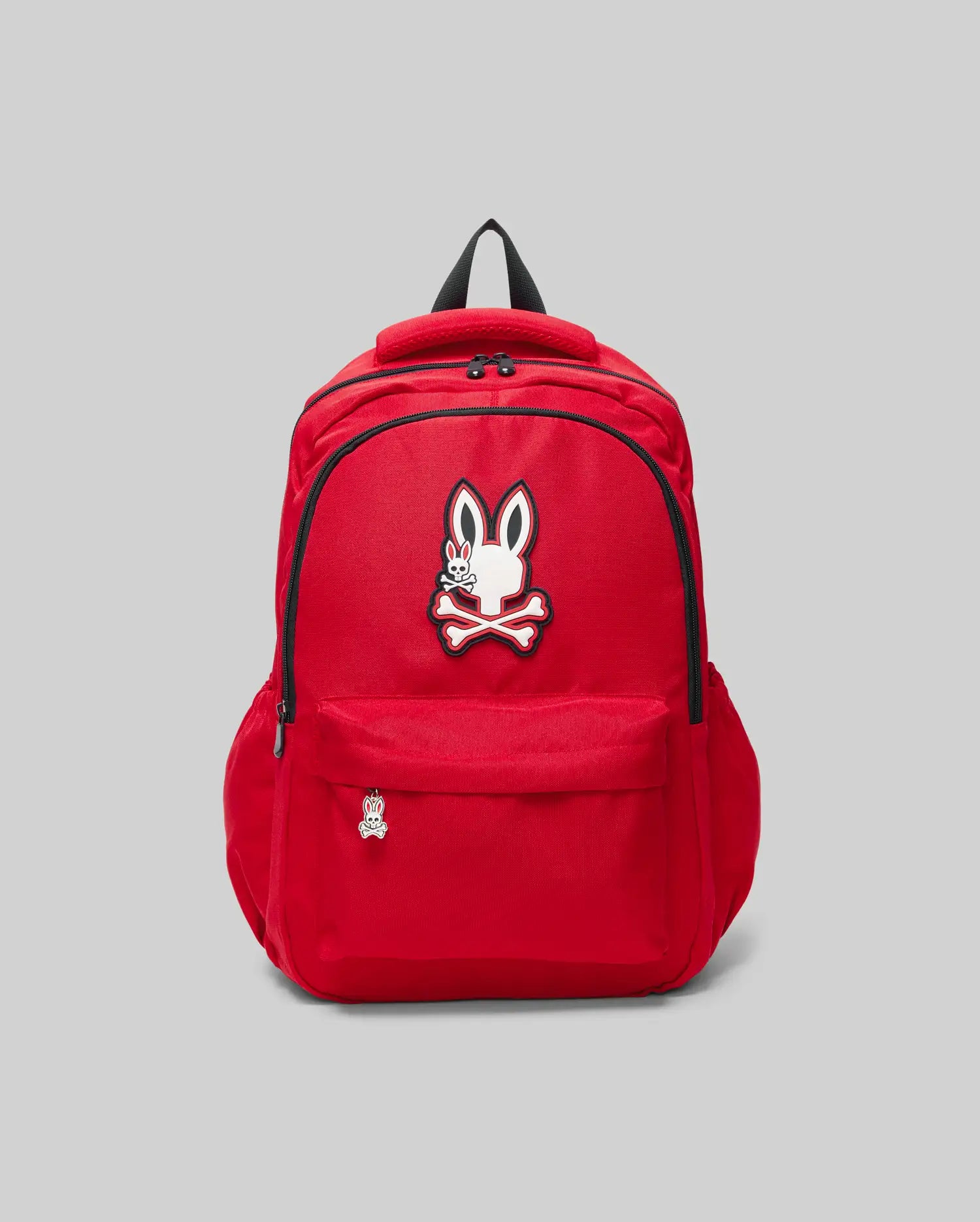 The KIDS PSYCHO BUNNY BACKPACK - KO2400004 by Psycho Bunny is a bright red canvas kids' bag with padded straps, front pocket, zipper, cartoon bunny with crossbones logo above the pocket, and a matching pin. Shown on a plain light gray background.