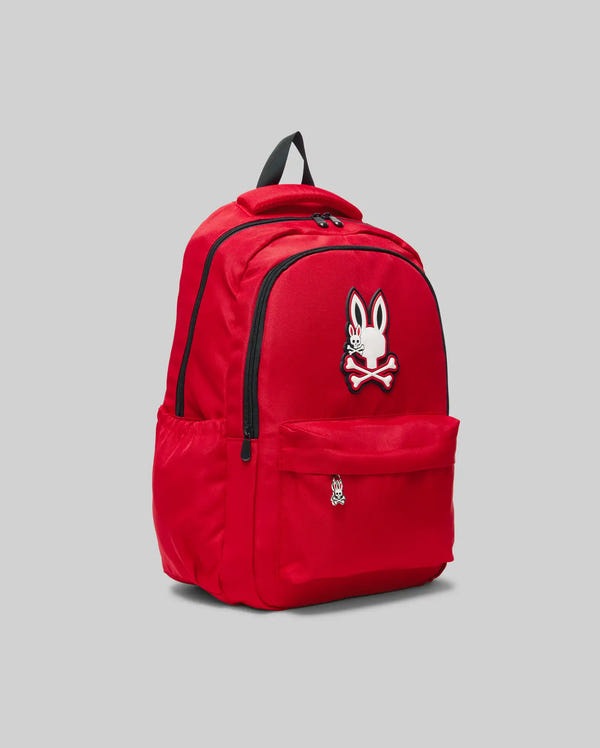 The KIDS PSYCHO BUNNY BACKPACK - KO2400004 by Psycho Bunny is a bright red canvas kids backpack with a cartoon bunny and crossbones patch, multiple compartments, side pockets, and padded straps, shown on a plain gray background.