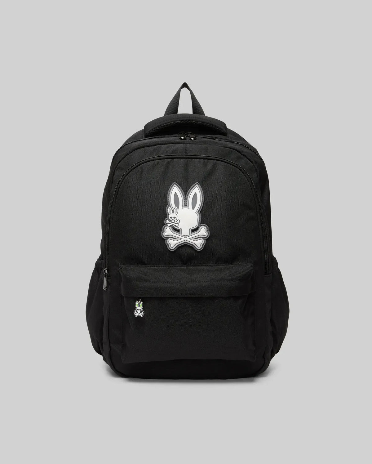 The Psycho Bunny KIDS PSYCHO BUNNY BACKPACK - KO2400004 displays a white cartoon bunny and crossbones emblem on the front pocket. Made from durable canvas with padded, adjustable straps, it is shown on a plain gray background.