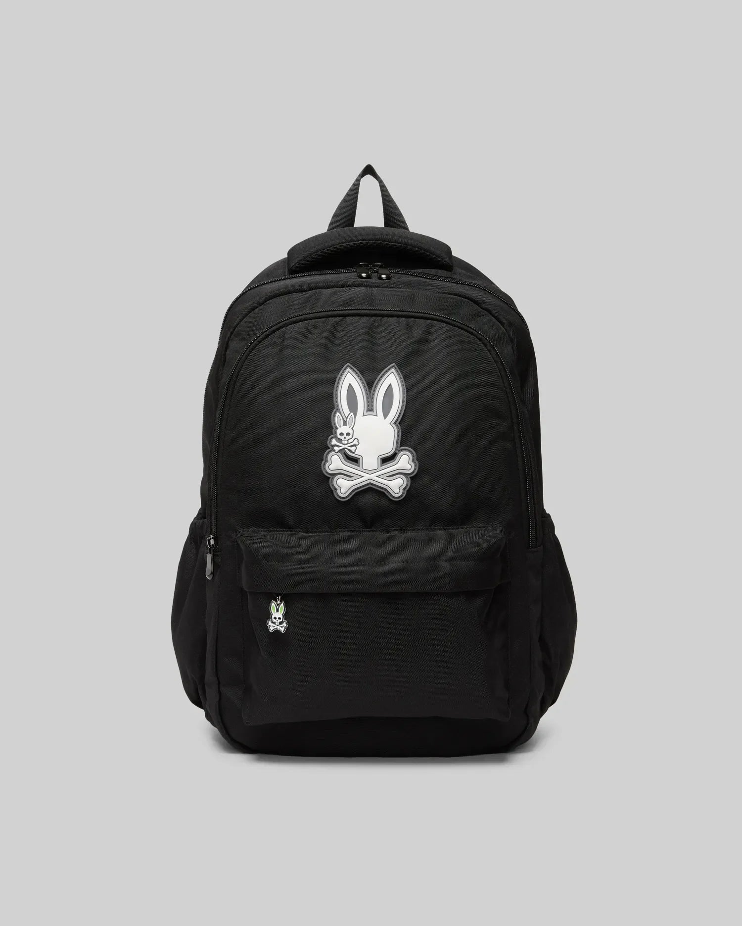 The Psycho Bunny KIDS PSYCHO BUNNY BACKPACK - KO2400004 displays a white cartoon bunny and crossbones emblem on the front pocket. Made from durable canvas with padded, adjustable straps, it is shown on a plain gray background.