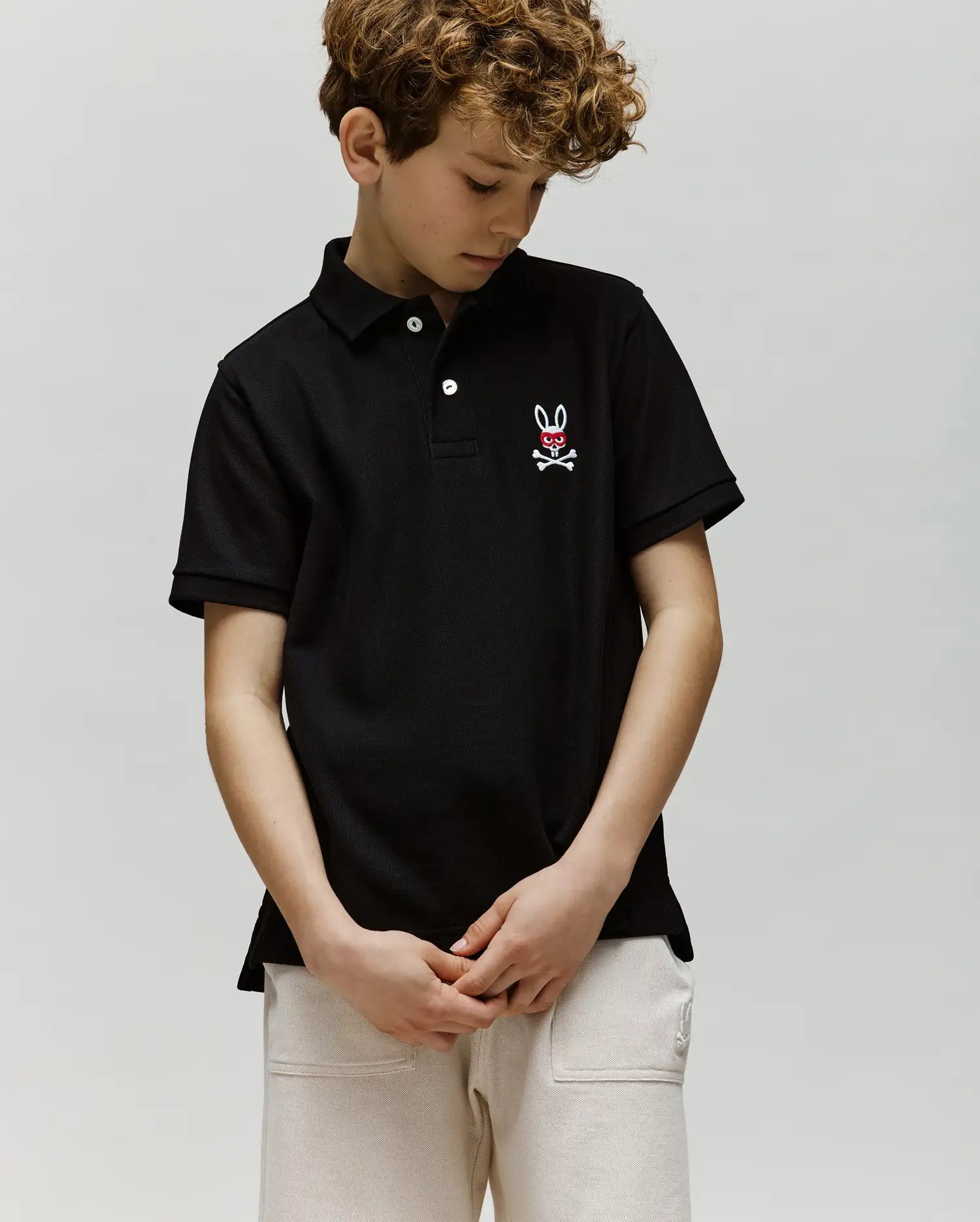 A boy with curly hair looks down wearing the Psycho Bunny KIDS MISCHIEF BUNNY PIQUE POLO (KT0100018), featuring soft Pima cotton, mother-of-pearl buttons, and a bunny and crossbones logo, paired with light-colored pants against a plain background.