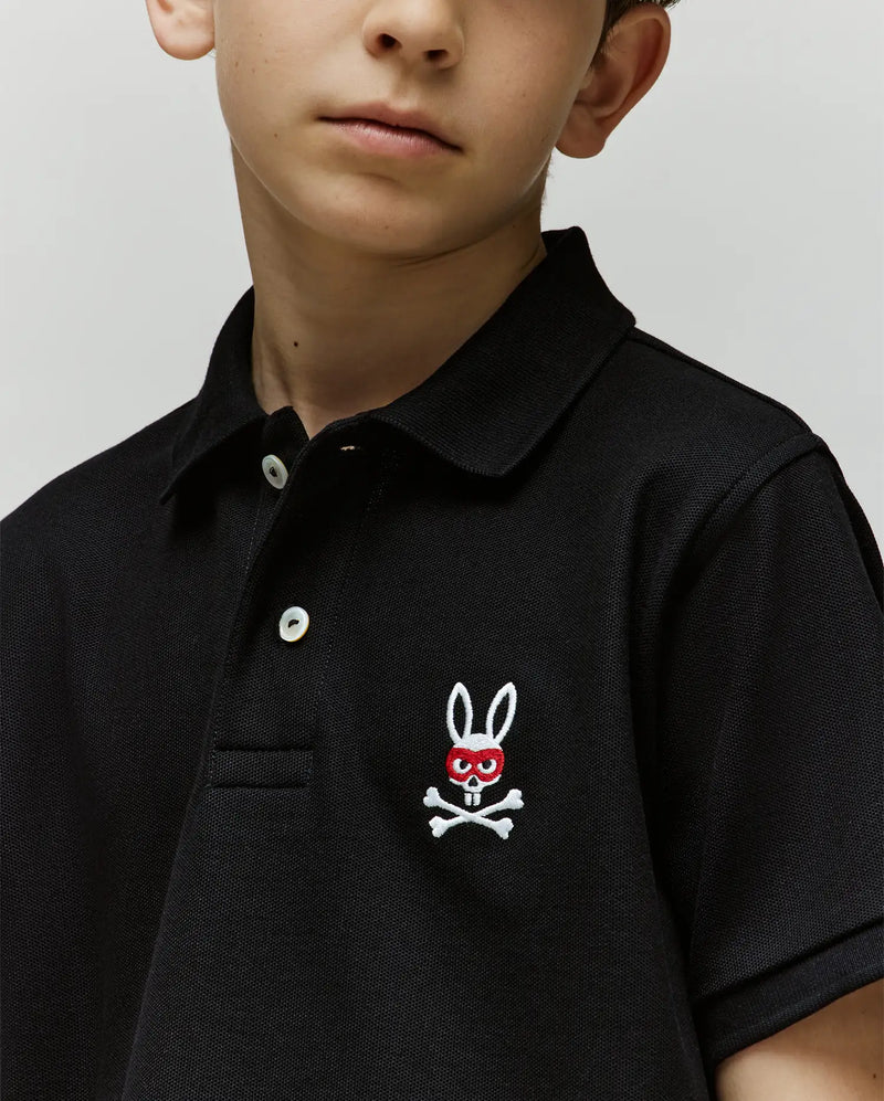 A young boy wears the Psycho Bunny KIDS MISCHIEF BUNNY PIQUE POLO (KT0100018), featuring soft Pima cotton, mother-of-pearl buttons, and a red-eyed bunny with crossbones patch. Image crops to highlight his shirt and part of his face.