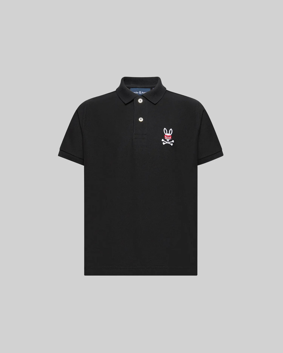 The Psycho Bunny KIDS MISCHIEF BUNNY PIQUE POLO (KT0100018) is a black, short-sleeve collared shirt with two mother-of-pearl buttons and a white and pink bunny skull logo on the left chest, shown on a light gray background.