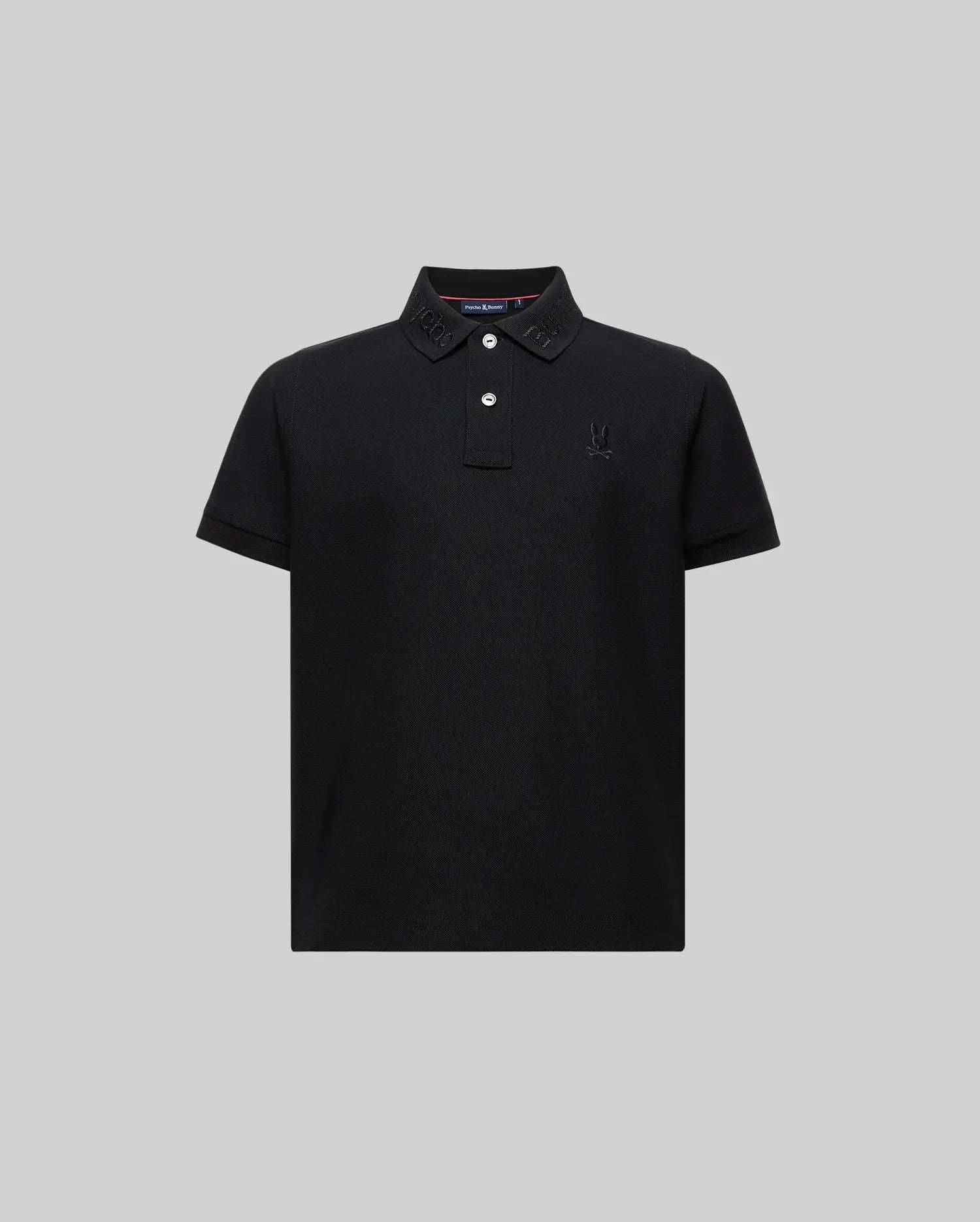 The Psycho Bunny KIDS CYRUS WORDMARK PIQUE POLO (KT0100026) is a soft Pima cotton, black short-sleeved polo with a collar, two white buttons, and subtle wordmark embroidery on the collar and chest, shown on a plain gray background.
