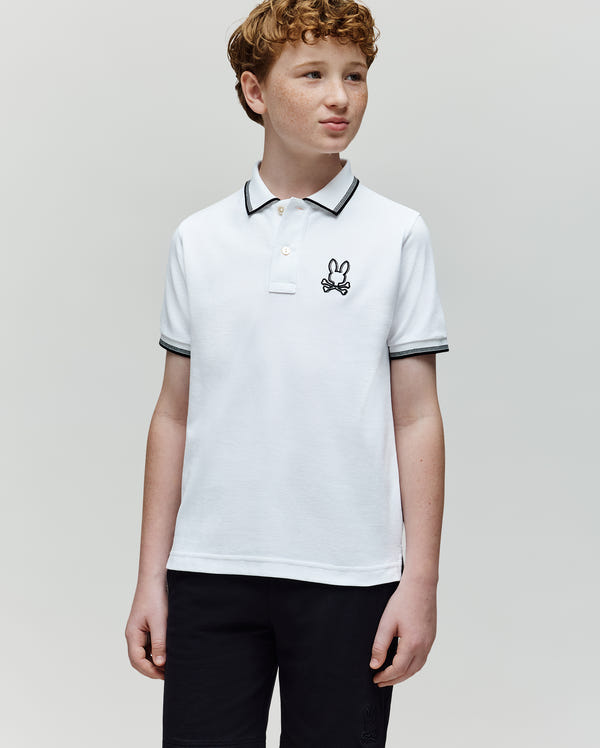 A curly-haired boy wears a white Psycho Bunny KIDS JUDE PIQUE POLO (KT0100027) with an embroidered bunny logo and black shorts, standing against a plain light gray background, looking slightly to his left.