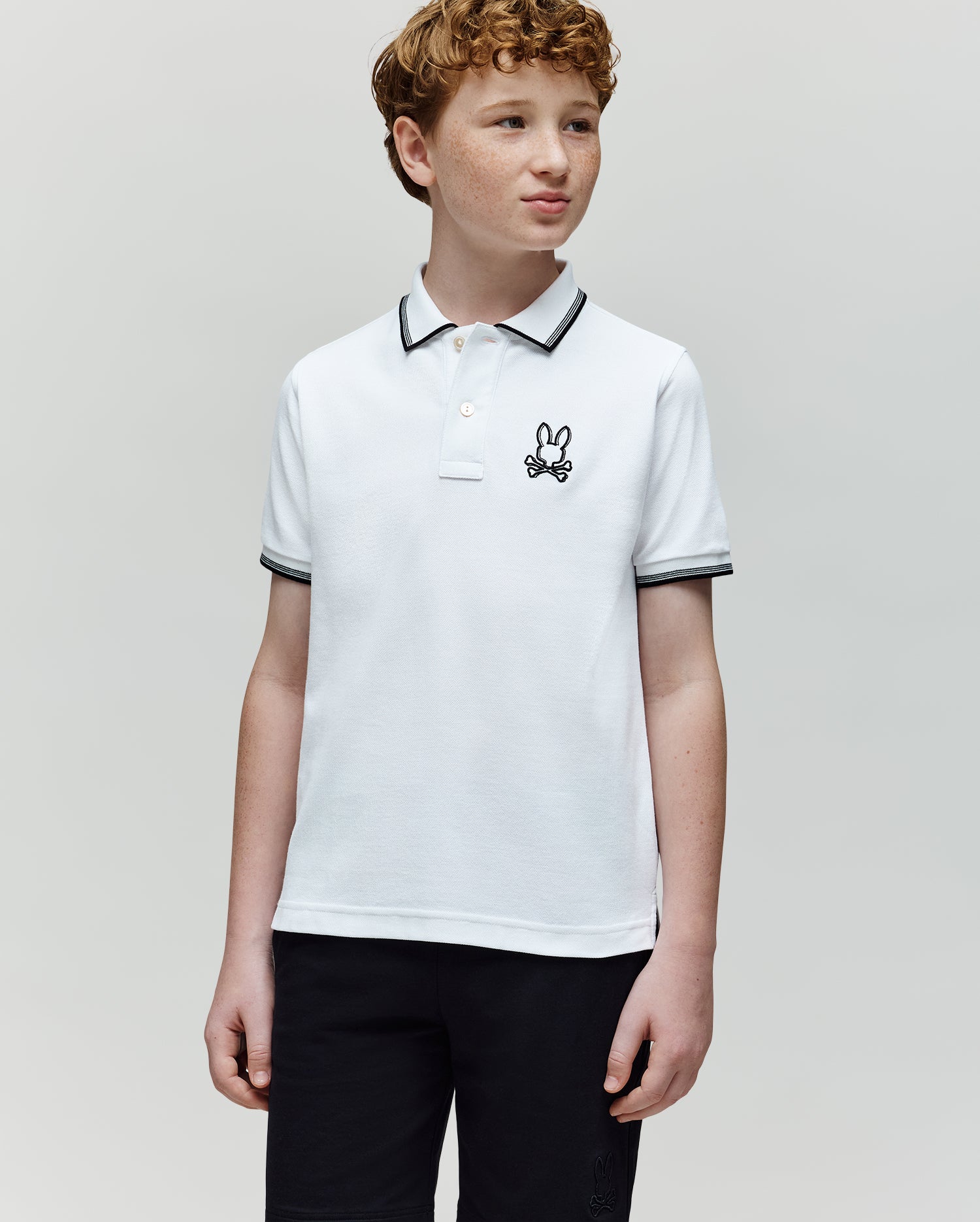 A curly-haired boy wears a white Psycho Bunny KIDS JUDE PIQUE POLO (KT0100027) with an embroidered bunny logo and black shorts, standing against a plain light gray background, looking slightly to his left.