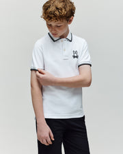 A boy with curly hair wears the Psycho Bunny KIDS JUDE PIQUE POLO (KT0100027) in white, made from soft Pima cotton with an embroidered Bunny logo, paired with black shorts, as he looks down and touches his arm against a plain background.