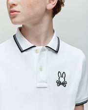 A young person wearing the Psycho Bunny KIDS JUDE PIQUE POLO (KT0100027), made from soft Pima cotton with black trim and an embroidered Bunny skull-and-crossbones logo on the chest, looks off to the side.