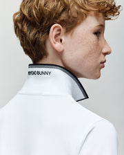 A young person with curly, light brown hair and freckles is seen in profile wearing the Psycho Bunny KIDS JUDE PIQUE POLO (KT0100027), featuring "PSYCHO BUNNY" text on the raised collar and an embroidered Bunny on premium Pima cotton. Plain light background.
