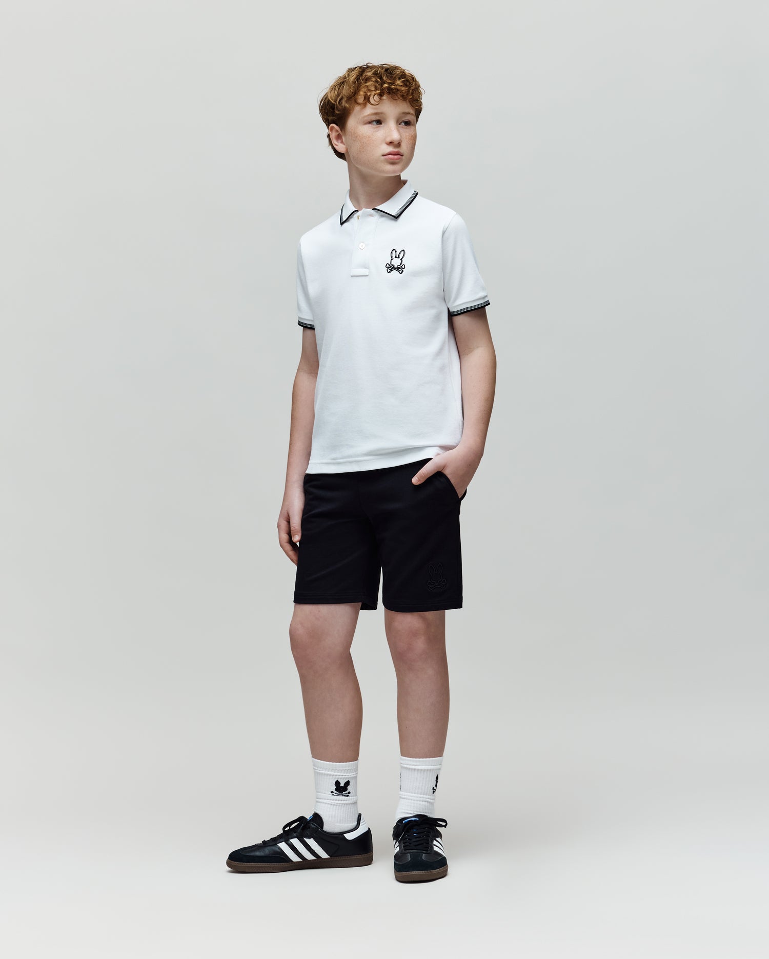 A young person with curly hair stands against a plain background, hands in pockets, wearing the Psycho Bunny KIDS JUDE PIQUE POLO - KT0100027 in white with an embroidered Bunny logo, black shorts, white socks, and black Adidas sneakers.