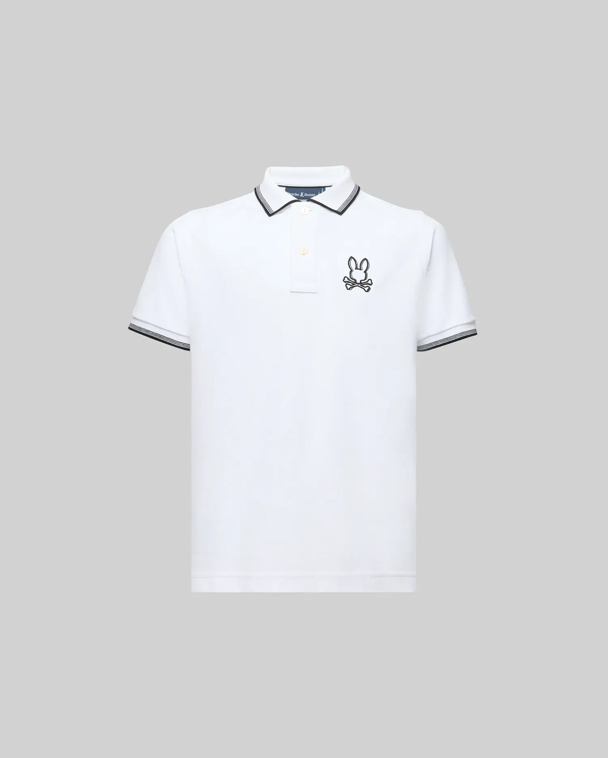 The Psycho Bunny KIDS JUDE PIQUE POLO (KT0100027) is a white Pima cotton short-sleeve polo with black trim on the collar and sleeve cuffs and an embroidered bunny logo on the left chest, shown against a plain gray background.